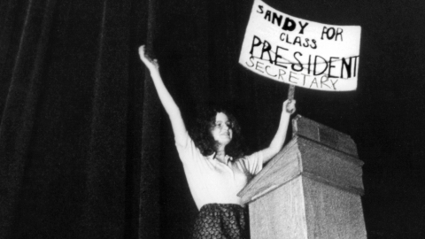 A young woman at a podium holding a sign that says Sandy for Class President, but president is crossed out and instead Secretary is written underneath