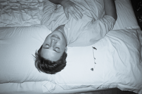 Young man ina white button down shirt. lying on a bed. White sheets with a rose next to him. 