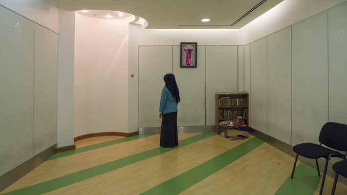 Photograph of a woman in a Muslim prayer space at an airport. She is facing toward Mecca with a yellow and green stripped floor and white walls. A corner of the room bears soft lighting.