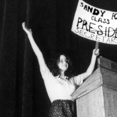 A young woman at a podium holding a sign that says Sandy for Class President, but president is crossed out with a big X and instead Secretary is written underneath. Black and White still from the film. 
