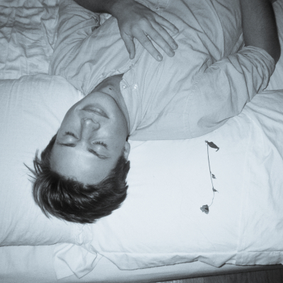 Young man ina white button down shirt. lying on a bed. White sheets with a rose next to him. 